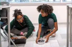 Two students sit on the ground. One holds a robotic device while the other types into a laptop.