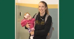 Chelsea Allen stands smiling in the school hall with her infant daughter in her arms.