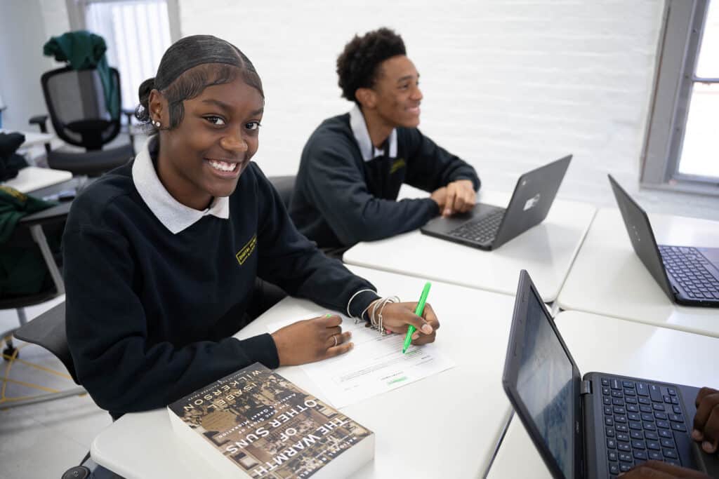 Two black high school students, one female and one male, sit at a table working, one on paper, one on a laptop.